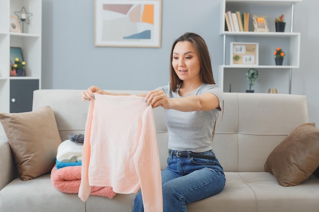 Young asian woman in casual clothes sitting on a couch with stack of clean laundry indoor at home happy and positive smiling