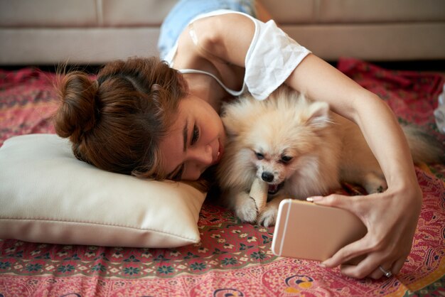 Young Asian woman lying on floor with small pet dog and taking selfies with smartphone