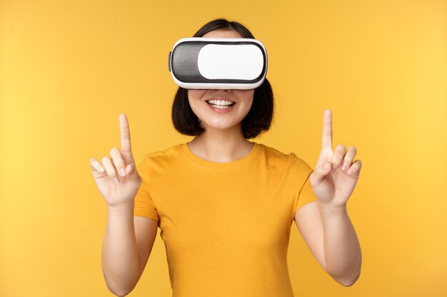 Young asian woman playing in VR using virtual reality glasses wearing yellow tshirt against studio background