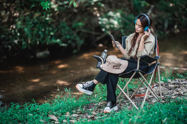 Young asian woman sit in a chair near the stream listening to music from tablet with wireless headphones happily while camping in the woods