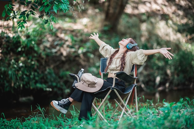 Free photo young asian woman sit in a chair near the stream listening to music from tablet with wireless headphones happily while camping in the woods