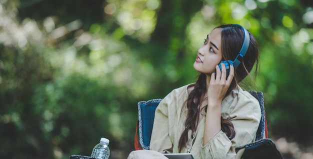 Young asian woman sit in a chair near the stream listening to music on wireless headphones and use tablet with happily while camping in the woods copy space