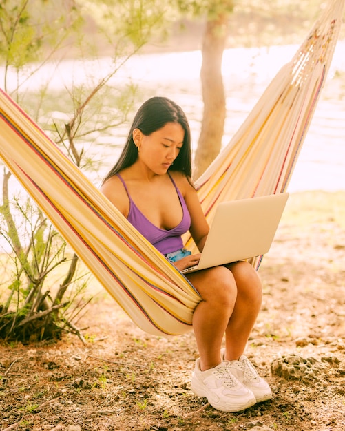 Free photo young asian woman sitting in hammock near river and freelancing on laptop