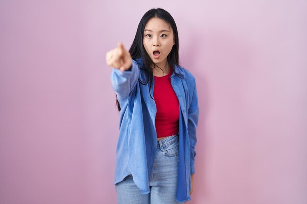 Young asian woman standing over pink background pointing with finger surprised ahead, open mouth amazed expression, something on the front