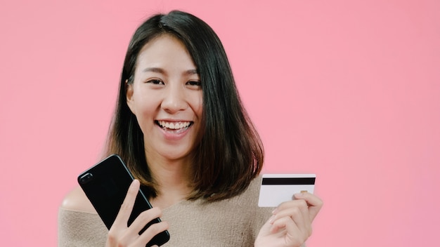 Young Asian woman using smartphone buying online shopping by credit card feeling happy smiling in casual clothing over pink background studio shot. 