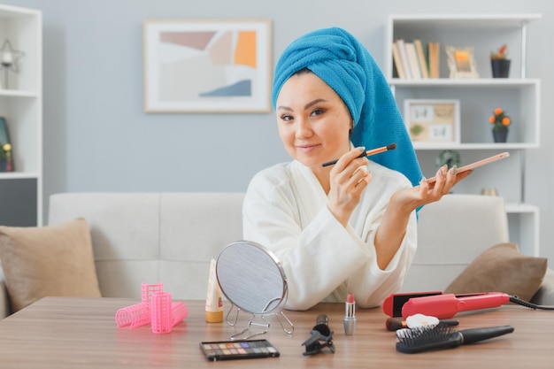 Free photo young asian woman with towel on her head sitting at the dressing table at home interior applying eye shadows holding palette smiling doing morning makeup routine beauty and facial cosmetics concept