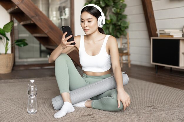 Young asian woman with water bottle listening music or podcast in headphones during fitness at home ...