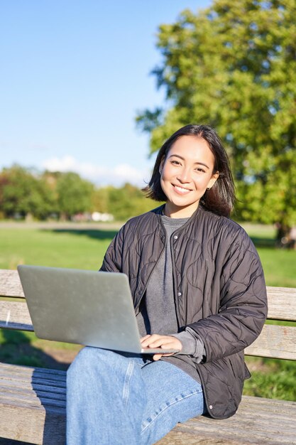 Young asian woman working remotely freelance girl sits in park with laptop doing her job from outdoo