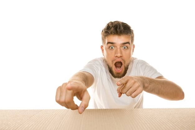 Young astonished man opening the biggest postal package isolated on white. Shocked male model on top of cardboard box looking inside. 