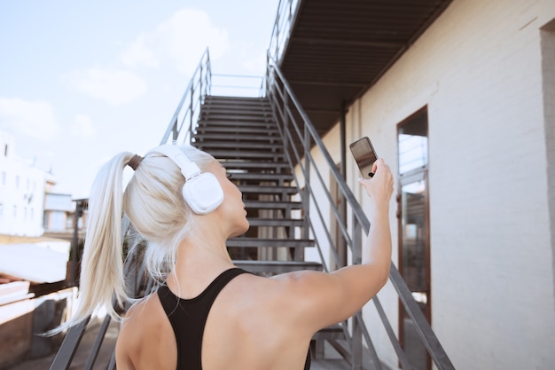 Free Photo a young athletic woman in shirt and white headphones working out listening to the music on a stairs outdoors