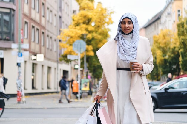 Young attractive Arabic woman in hijab walking through city street with shopping bags and coffee