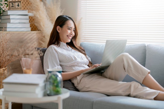 Young attractive asian female working at home hand use laptop checking search schedule working process planning report with leisure relax on sofa in living room with morning light peaceful moment