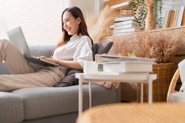 Young attractive asian female working at home hand use laptop checking search schedule working process planning report with leisure relax on sofa in living room with morning light peaceful moment