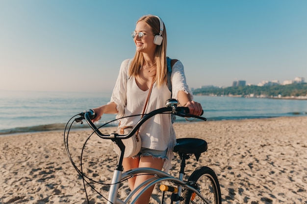 Young attractive blond smiling woman walking on beach with bicycle in headphones