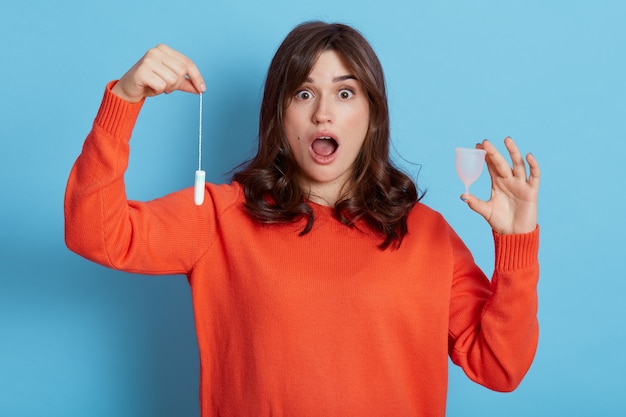 Young attractive brunette female wearing casual clothing holding tampon and menstrual cup in hands