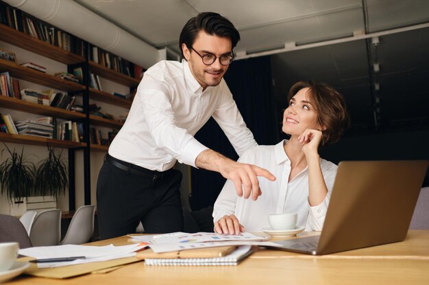 Young attractive business colleagues happily working on new project with laptop and papers in modern office