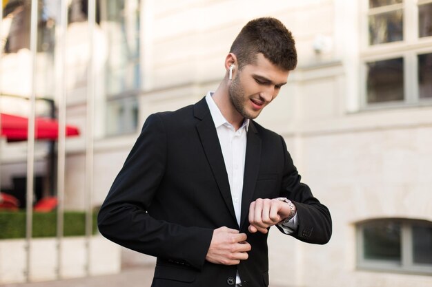 Young attractive businessman in black suit and white shirt with wireless earphones dreamily looking time on hand watch while spending time outdoor