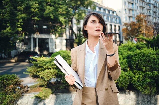 Young attractive businesswoman in beige suit with laptop thoughtfully recording voice message on cellphone on city street