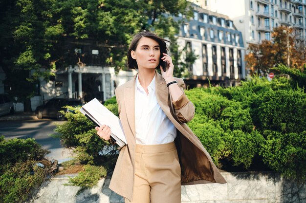 Young attractive businesswoman in beige suit with laptop thoughtfully talking on cellphone on city street