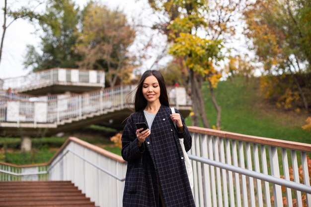 Young attractive casual woman in coat happily using cellphone while walking in city park