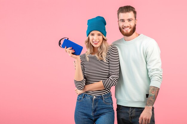 Young attractive couple listening to music on wireless speaker wearing cool stylish outfit smiling happy positive mood posing on pink background 