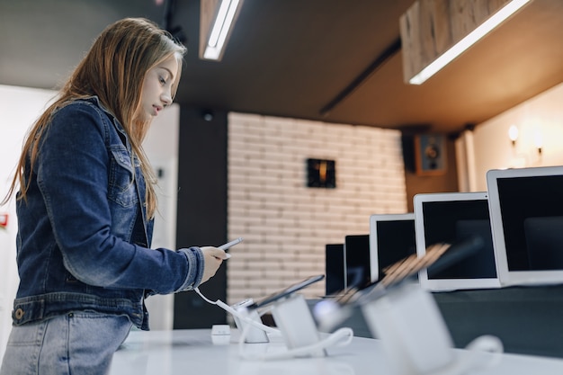 Young attractive girl in electronics store stands at desk and tests phone