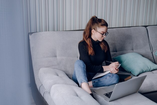 Young attractive girl at home working with laptop on the couch. comfort and coziness while at home. home office and work from home. remote online employment.