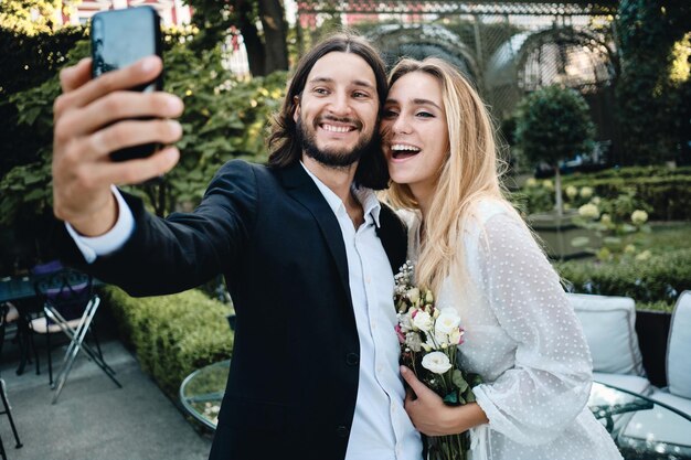 Young attractive groom and bride joyfully taking selfie on cellphone together in beautiful garden of restaurant