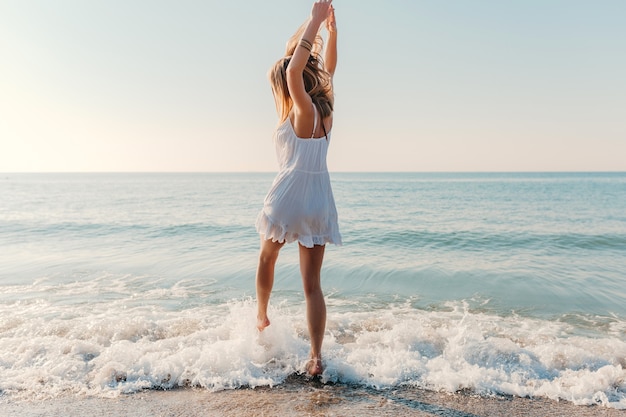 Young attractive happy woman dancing turning around by sea beach sunny summer fashion style in white dress vacation