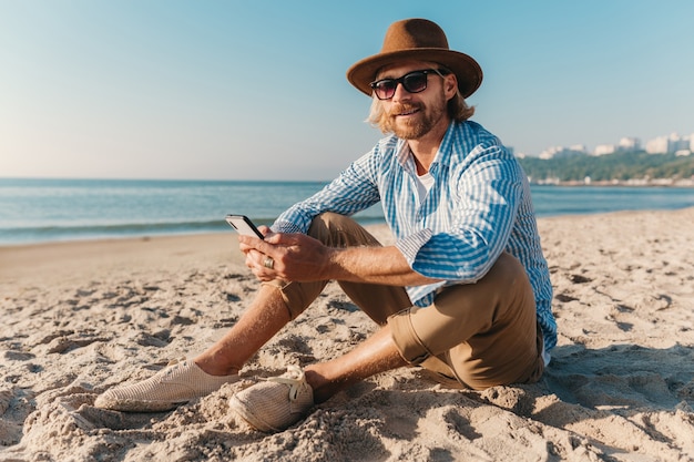 Young attractive hipster man sitting on beach by sea on summer vacation