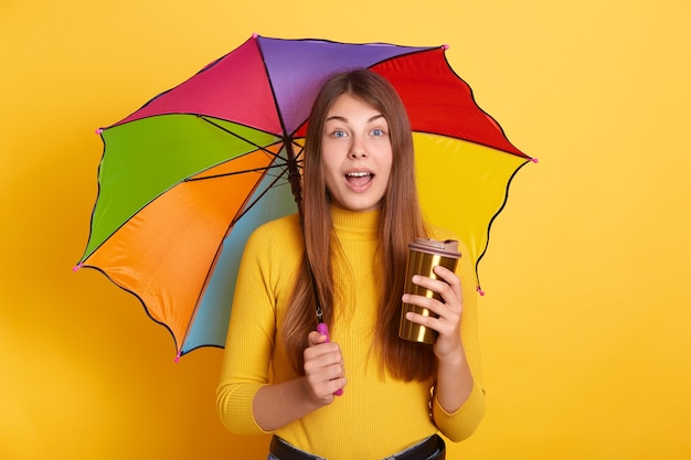 Free photo young attractive lady with astonished facial expression posing with multicolored umbrella and coffee to go, stands with opened mouth