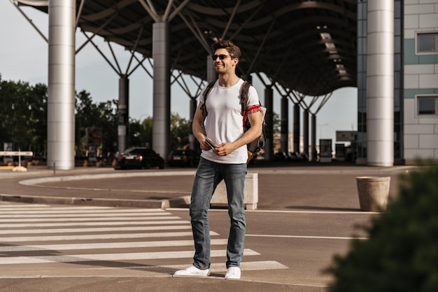 Young attractive man in jeans white tee and sunglasses holds passport and backpack Brunette guy in good mood moves in city