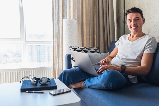 Young attractive man sitting on sofa at home working on laptop online, using internet