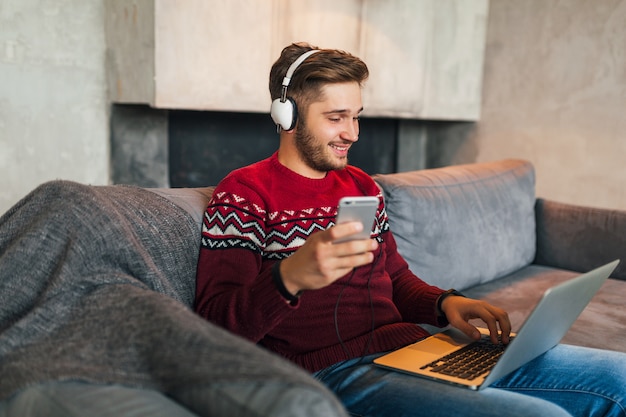 Free photo young attractive man on sofa at home in winter with smartphone in headphones, listening to music, wearing red knitted sweater, working on laptop, freelancer, smiling, happy, positive, typing