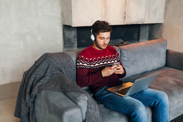 Free photo young attractive man on sofa at home in winter with smartphone in headphones, listening to music, wearing red knitted sweater, working on laptop, freelancer