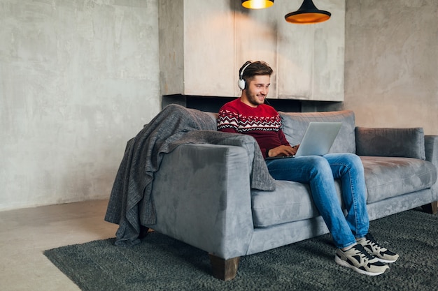 Free photo young attractive man on sofa at home in winter with smartphone in headphones, listening to music, wearing red knitted sweater, working on laptop, freelancer