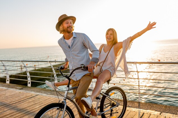 Young attractive man and woman traveling on bicycles, romantic couple on summer vacation by the sea on sunset, boho hipster style outfit, friends having fun together