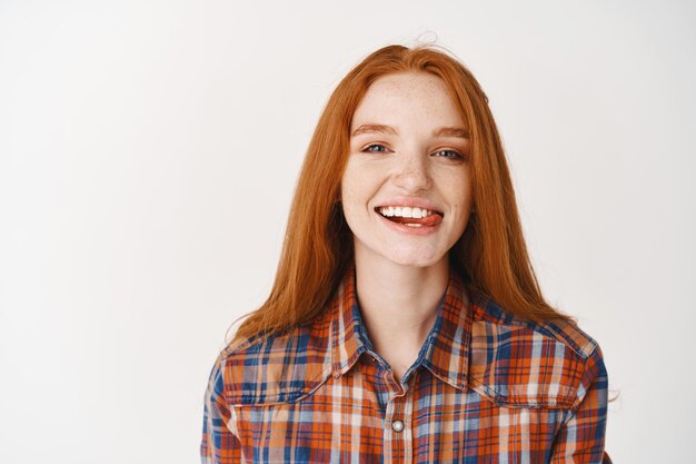 Young attractive redhead girl showing tongue, smiling with white teeth, looking carefree at front, standing over white wall