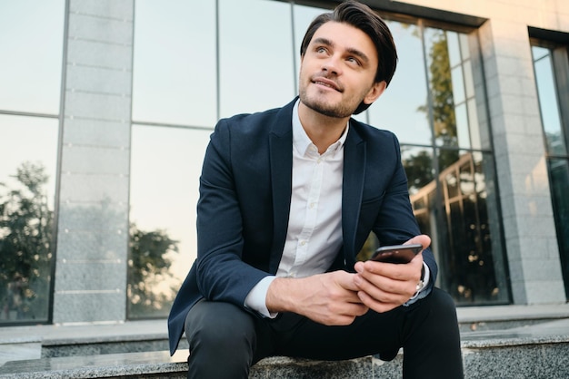 Young attractive smiling bearded brunette man in white shirt and classic suit dreamily looking aside holding cellphone in hand with glass building on background