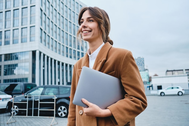 Free Photo young attractive smiling businesswoman in coat with laptop happily looking away while walking through city street