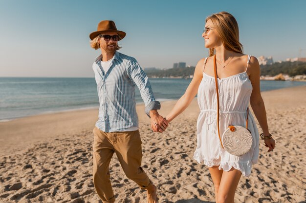 Young attractive smiling happy man in hat and blond woman in white dress running together on beach