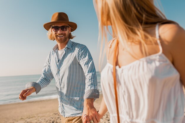 Young attractive smiling happy man in hat and blond woman in white dress running together on beach