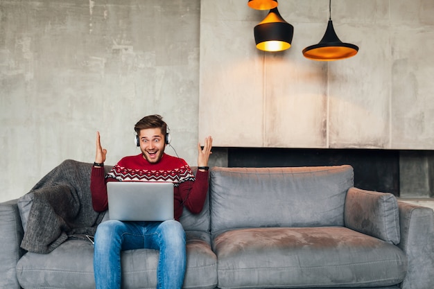 Free photo young attractive smiling man sitting on sofa at home in winter with surprised face expression, wearing red knitted sweater, working on laptop, freelancer, emotional, screaming, listening to headphones