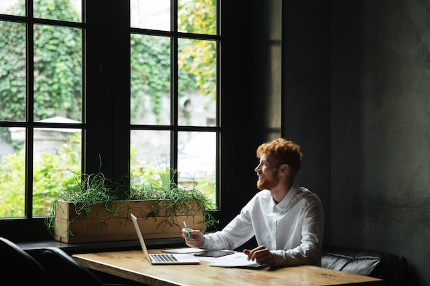 Free Photo young attractive smiling redhead bearded business man looking at window while sitting at workplace