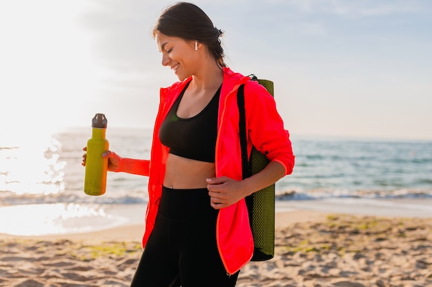 Free photo young attractive smiling woman doing sports in morning sunrise on sea beach holding yoga mat and bottle of water, healthy lifestyle, listening to music on earphones, wearing pink windbreaker jacket