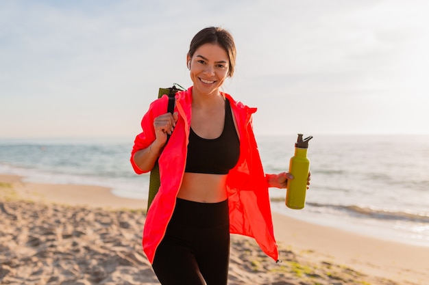 Free photo young attractive smiling woman doing sports in morning sunrise on sea beach holding yoga mat and bottle of water, healthy lifestyle, listening to music on earphones, wearing pink windbreaker jacket