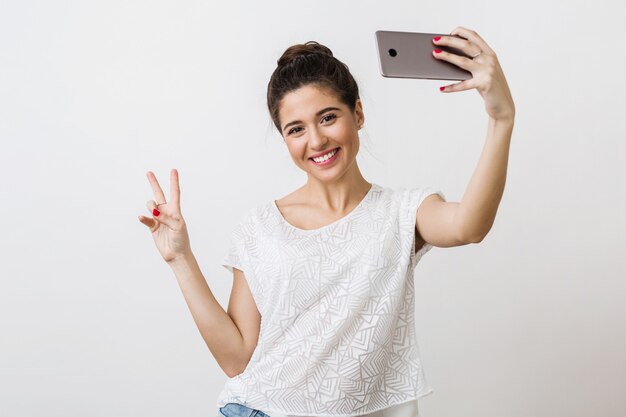 Young attractive smiling woman in white blouse holding smart phone, showing peace sign, positive emotion, making selfie photo, 