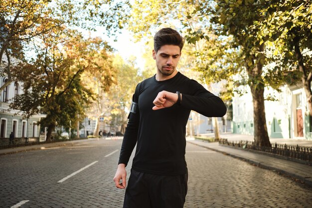 Young attractive sporty man thoughtfully checking his watch after morning run on city street