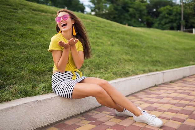 Free photo young attractive stylish smiling woman having fun in city park, positive, emotional, wearing yellow top, striped mini skirt, pink sunglasses, white sneakers, summer style fashion trend, long legs