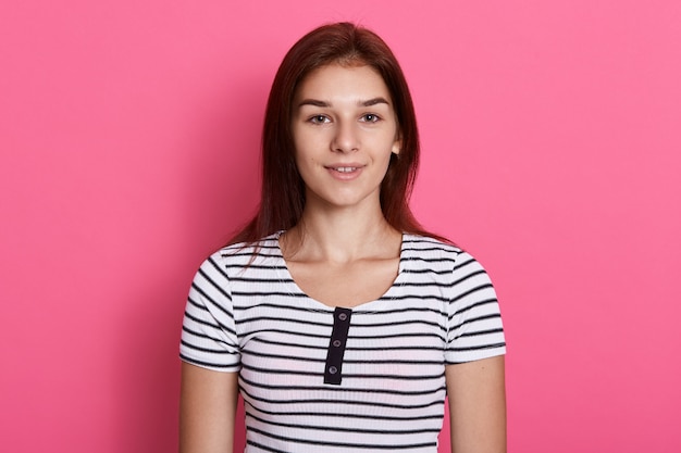 Young attractive teenage girl wearing striped casual t shirt, posing against pink wall,  with charming smile, looks happy.